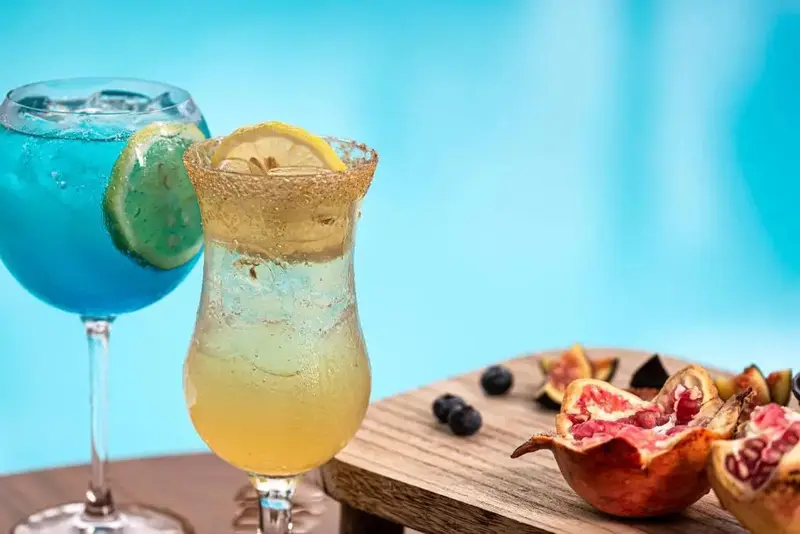 Deux cocktails colorés et des fruits frais sont présentés sur une planche en bois devant un flou bleu.