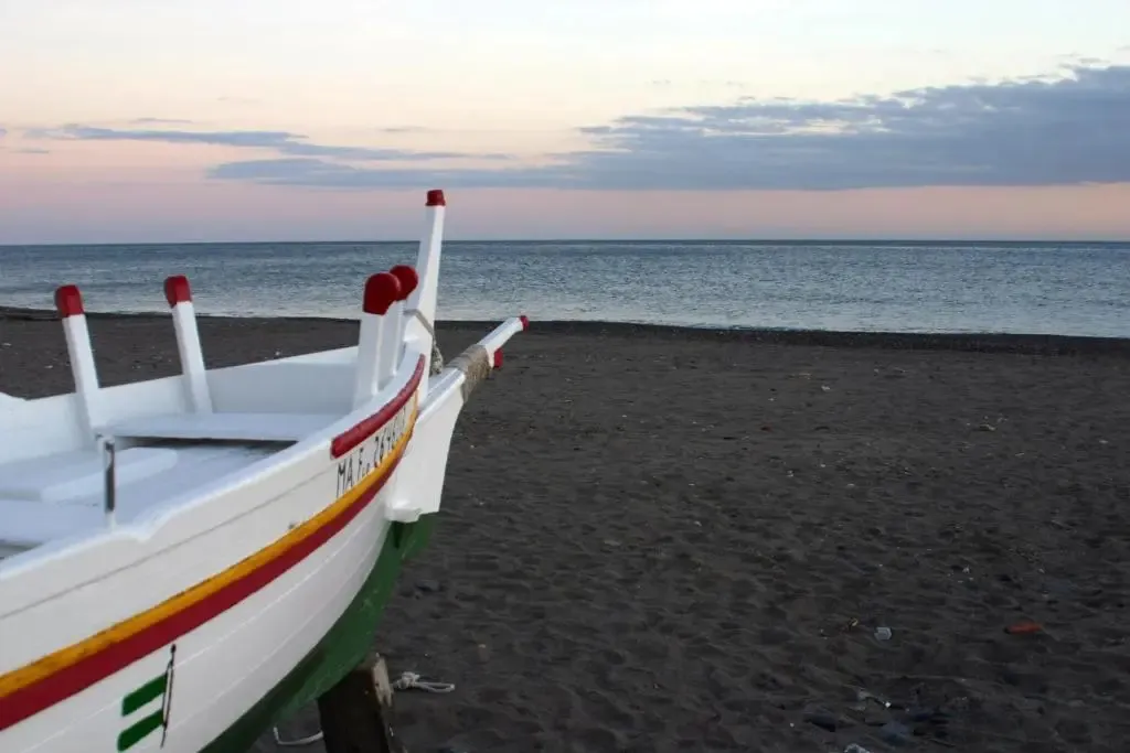 Un bateau de pêche coloré repose sur une plage de sable sombre au coucher du soleil.