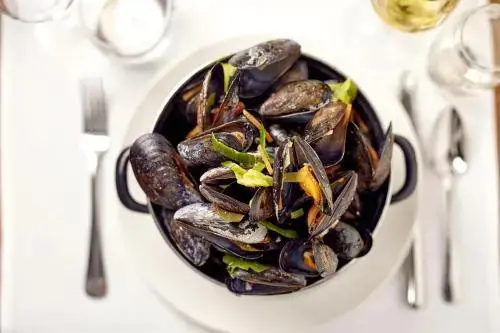 Un plat de moules cuisinées est présenté dans une cocotte avec des couverts et des verres.