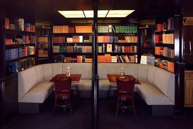 Deux tables en bois sont disposées face à des banquettes grises dans une pièce bordée d'étagères remplies de livres.