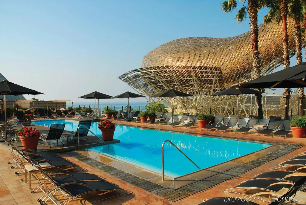 Une piscine bordée de transats sous des parasols, avec une structure architecturale en arrière-plan.