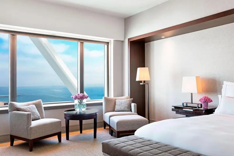 Une chambre lumineuse avec un lit et deux fauteuils près d'une grande fenêtre donnant sur la mer.