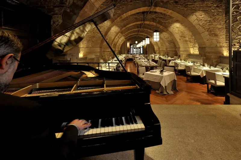 Un pianiste joue dans une salle voûtée éclairée, avec des tables dressées pour un repas.