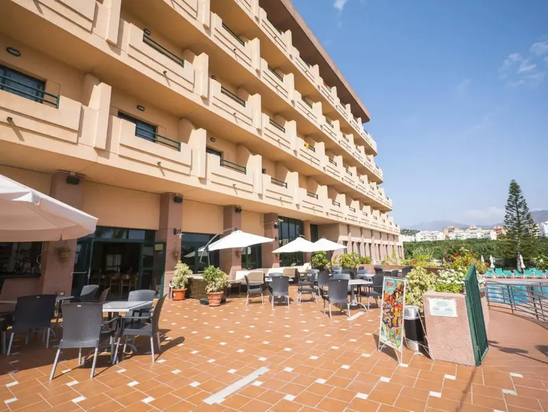Terrasse ombragée avec tables et chaises devant un bâtiment beige à balcons.