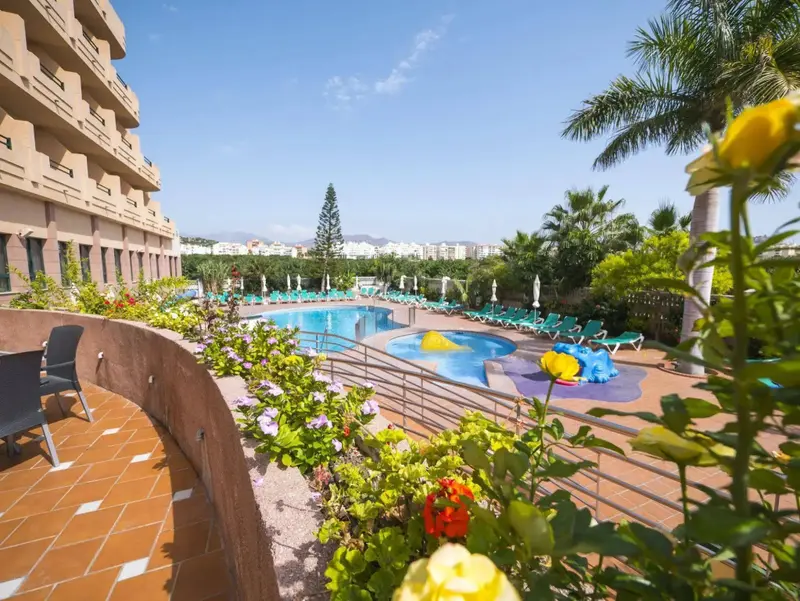 Une piscine ensoleillée avec des transats et des palmiers, devant un bâtiment à plusieurs étages.