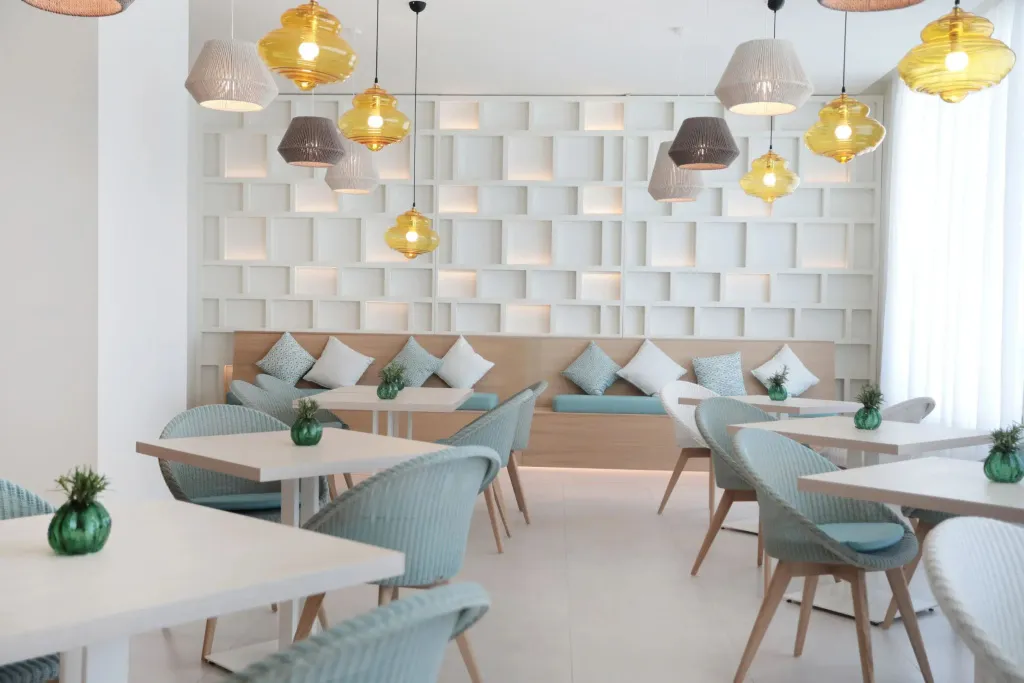 Une salle lumineuse avec des tables et des chaises turquoise, des suspensions colorées et un mur texturé.