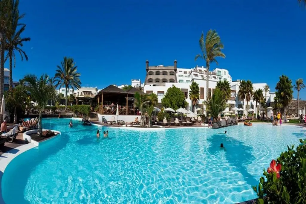 Une grande piscine turquoise avec des transats et des palmiers sous un ciel bleu.