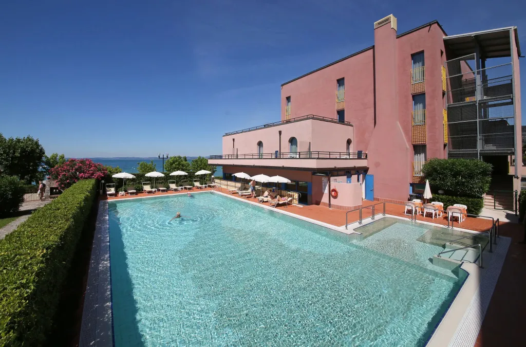 Une piscine rectangulaire turquoise bordée de transats et de parasols, devant un bâtiment rose.