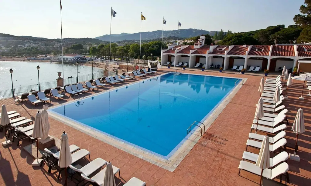 Une piscine rectangulaire bordée de transats sous des parasols, avec une vue sur la mer et des collines verdoyantes.