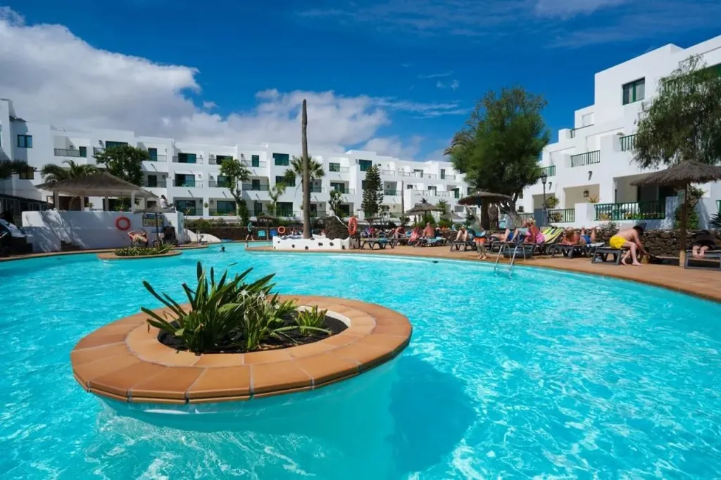 Une piscine turquoise au premier plan, entourée de transats et de bâtiments blancs.