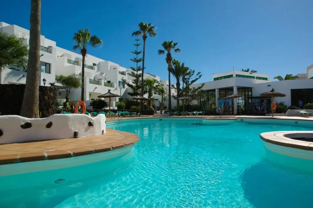 Piscine turquoise entourée d'un bâtiment blanc et de palmiers sous un ciel bleu.