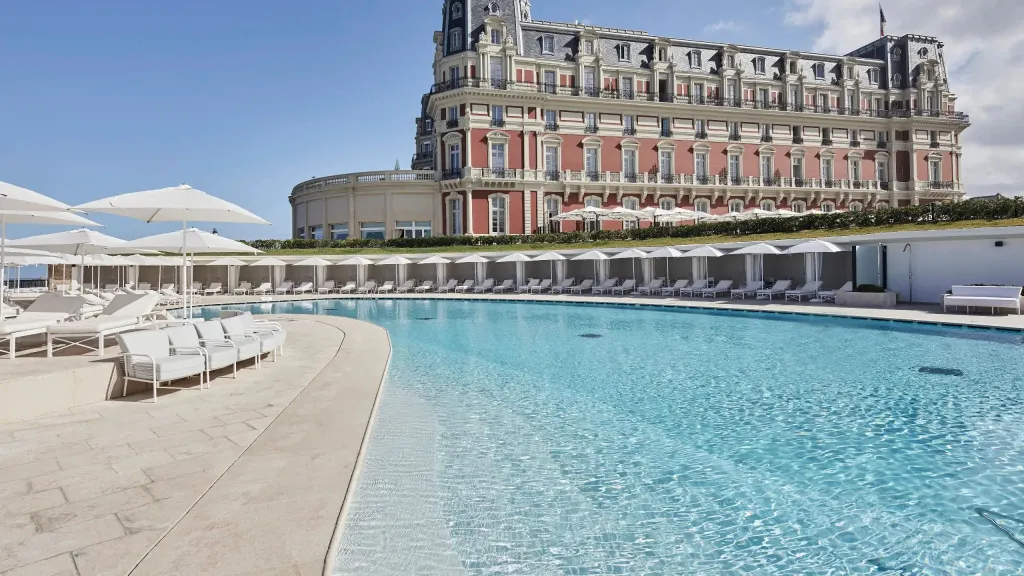 Une piscine bleue bordée de transats blancs devant un bâtiment imposant.