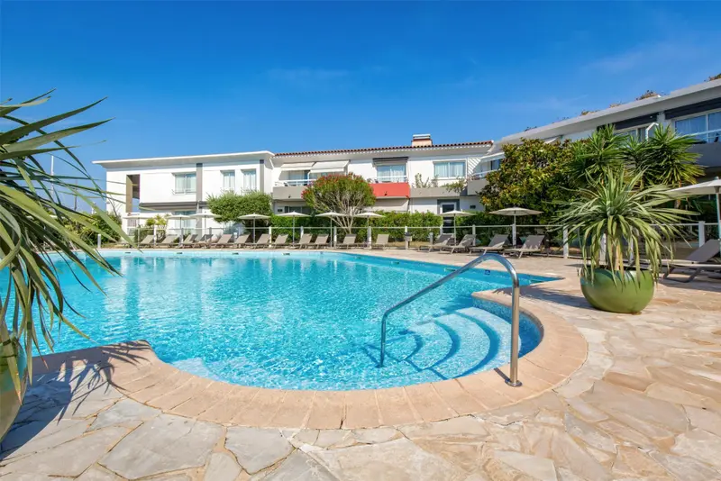 Une piscine rectangulaire avec des transats et des parasols, devant un bâtiment blanc à plusieurs étages.
