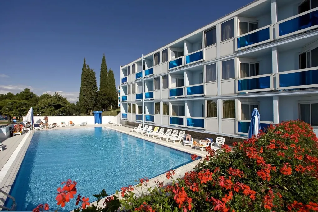 Une piscine turquoise bordée de transats et de fleurs, devant un bâtiment à balcons bleus.