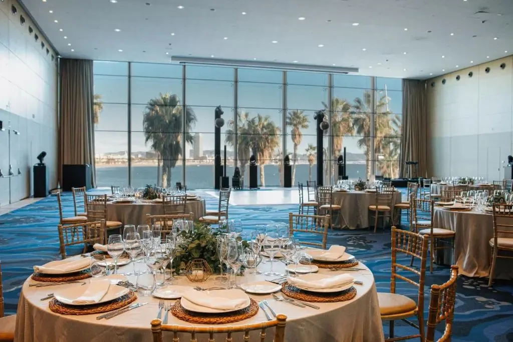 Une salle lumineuse est mise en scène pour un banquet avec des tables rondes nappées et décorées.