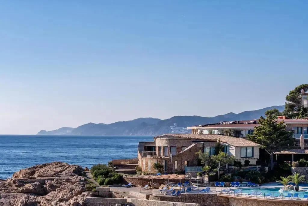Vue d'un établissement sur une côte rocheuse ensoleillée avec une piscine et la mer en arrière-plan.