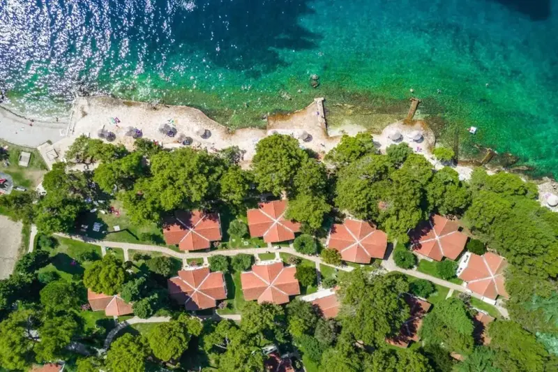 Vue aérienne d'un ensemble de bâtiments au toit rouge entourés de végétation, bordant une plage et une mer turquoise.