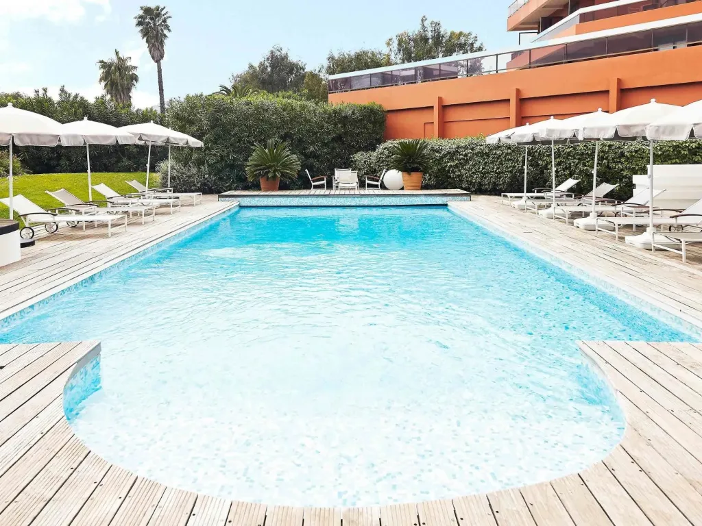 Une piscine rectangulaire entourée d'une terrasse en bois avec des transats et des parasols blancs.