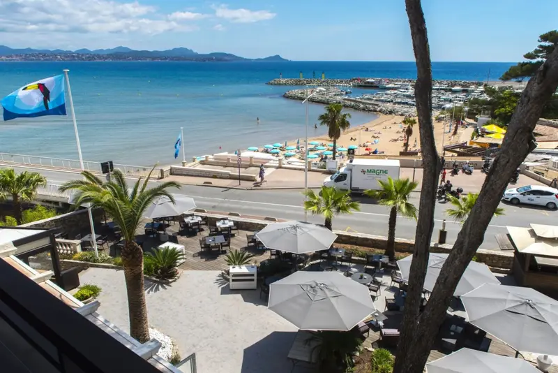 Terrasse ombragée avec tables et parasols donnant sur la mer et une plage.