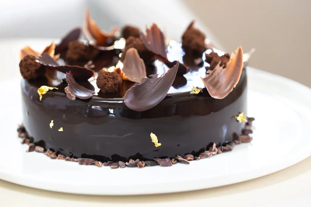 Un gâteau au chocolat décoré de copeaux et de feuilles de chocolat est présenté sur une assiette blanche.