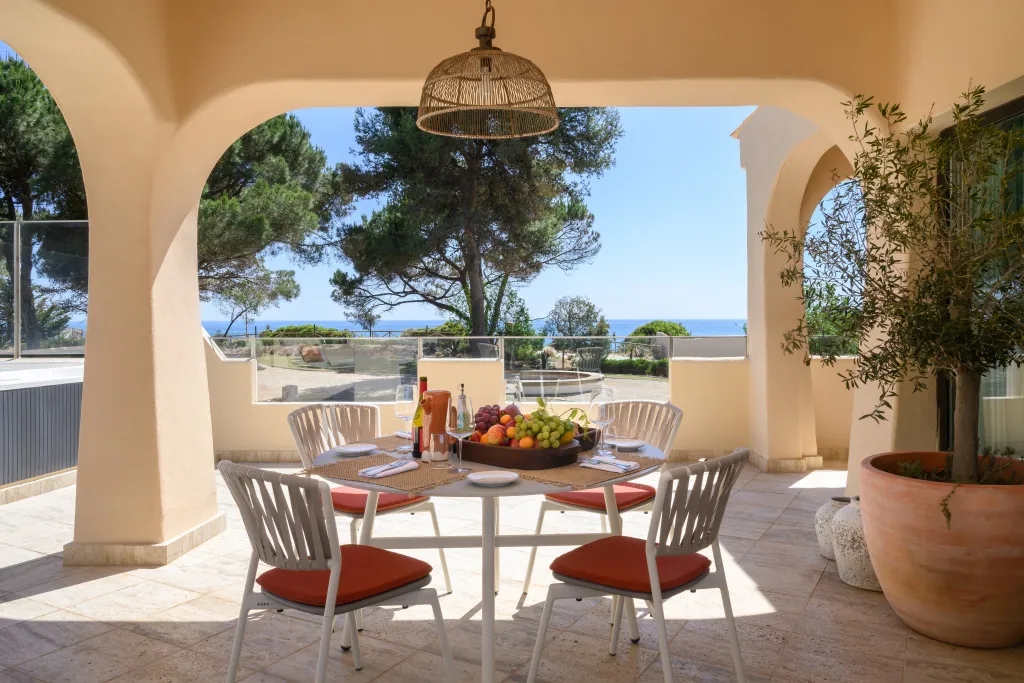 Une table ronde est dressée sur une terrasse ombragée avec vue sur la mer.