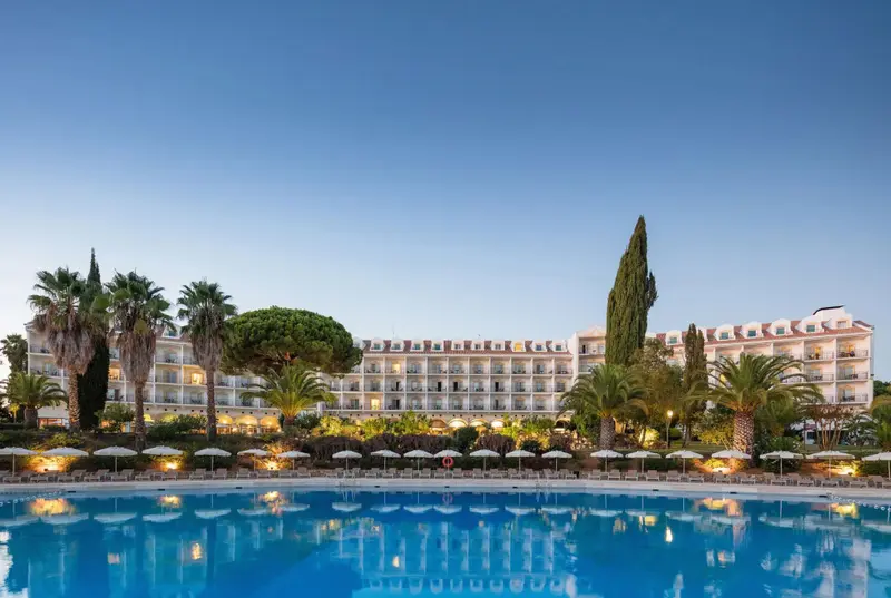 Bâtiment blanc à plusieurs étages avec piscine au premier plan et végétation tropicale.
