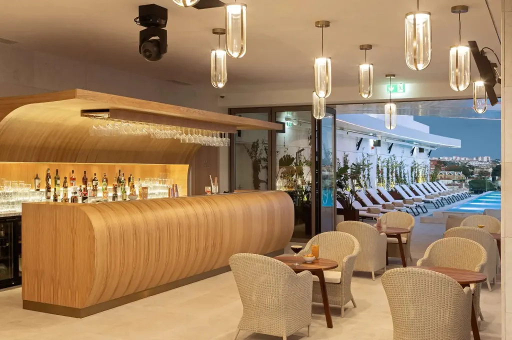 Un bar moderne avec un comptoir en bois clair et des tabourets, donnant sur une piscine et des transats.