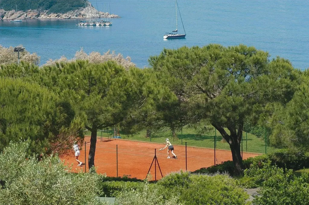 Deux personnes jouent au tennis sur un court en terre battue, avec une vue sur la mer en arrière-plan.