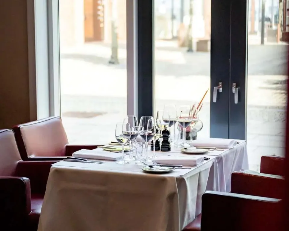Une table dressée avec de la vaisselle et des verres devant une grande fenêtre.