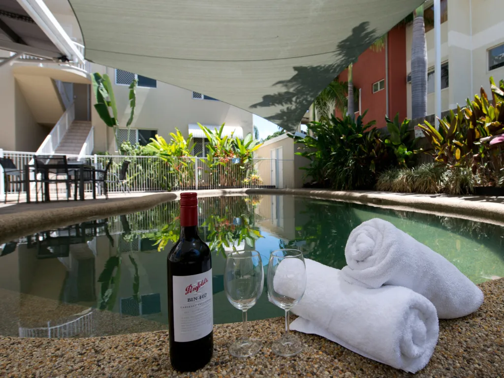 Une bouteille de vin et deux verres sont posés près d'une piscine ombragée par une voile.