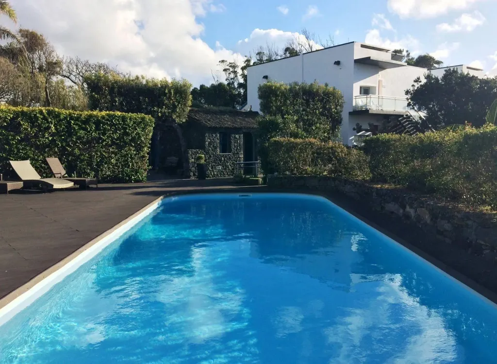 Une piscine rectangulaire bordée d'une terrasse en bois, avec un bâtiment blanc et une végétation luxuriante en arrière-plan.
