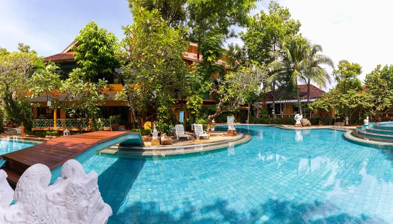 Une piscine turquoise sinueuse avec un pont en bois et des chaises longues à proximité d'un bâtiment tropical.