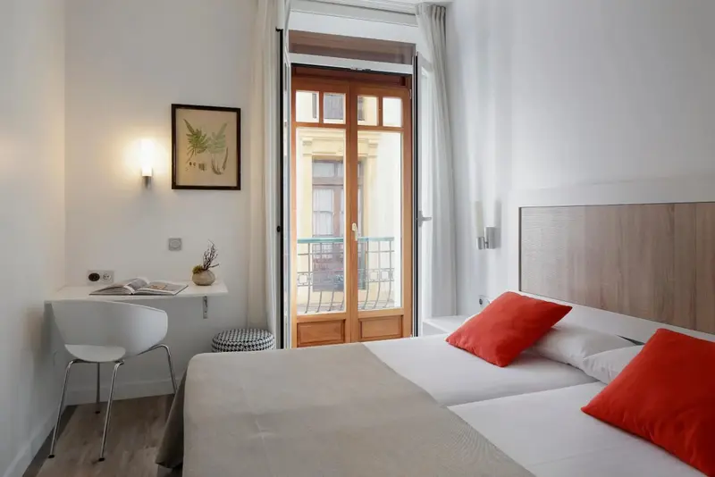 Une chambre lumineuse avec un lit double et un bureau près d'une porte-fenêtre ouverte sur un balcon.