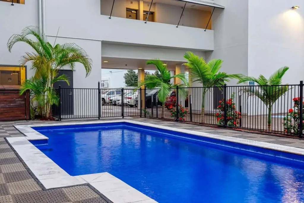 Une piscine rectangulaire bleue entourée d'une terrasse carrelée et de végétation tropicale.
