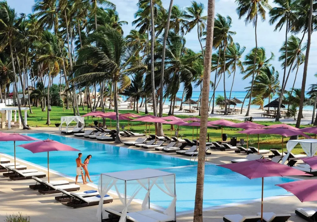 Une piscine bordée de transats et de parasols, avec une plage et des palmiers en arrière-plan.
