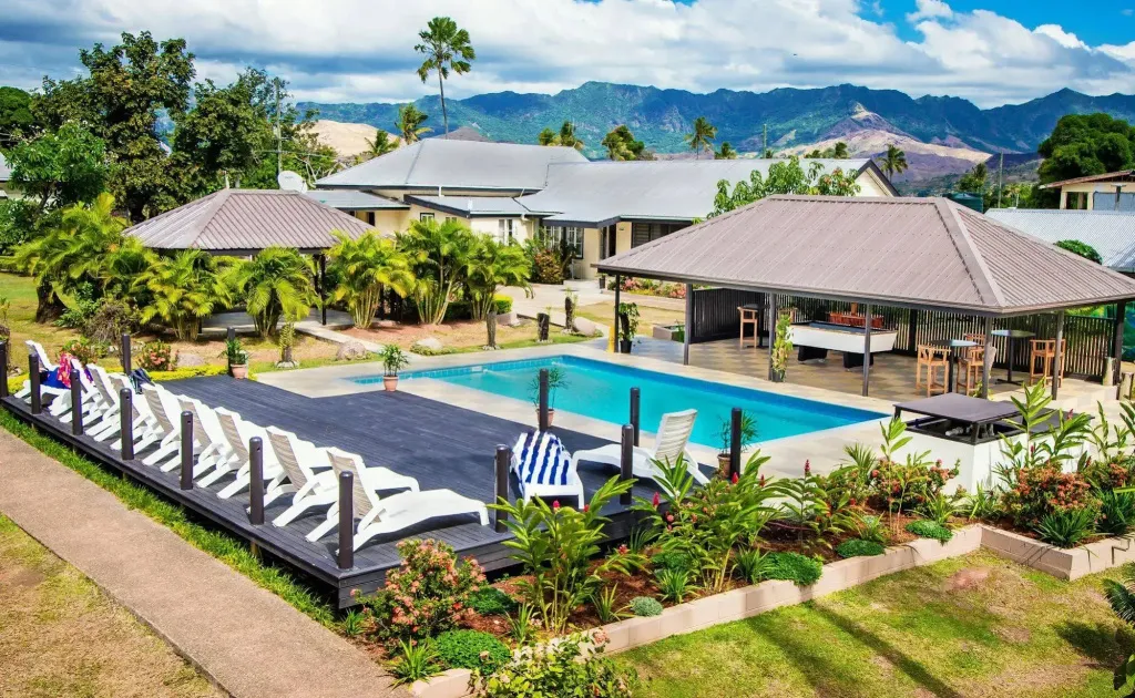 Une piscine bordée de transats dans un jardin tropical, avec des bâtiments en arrière-plan.