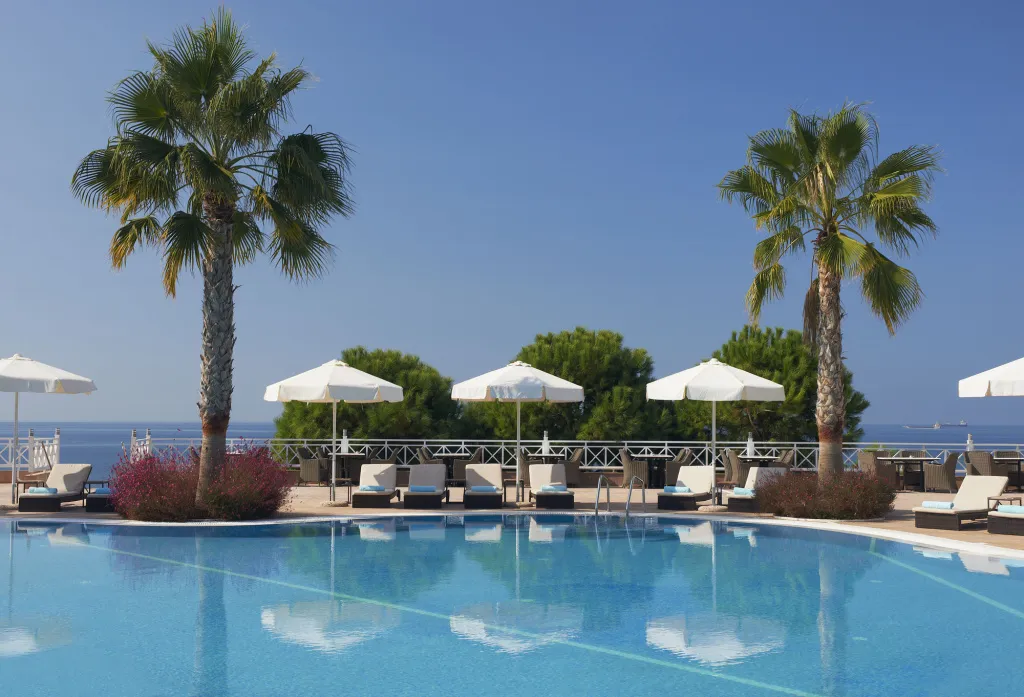 Transats blancs disposés au bord d'une piscine turquoise sous des parasols, avec une vue sur la mer.