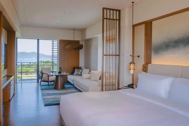 Une chambre lumineuse avec un grand lit et un espace salon donnant sur une vue verdoyante.