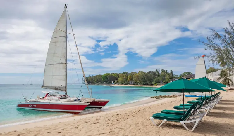 Une plage de sable blanc avec des transats verts et un catamaran rouge amarré dans l'eau turquoise.