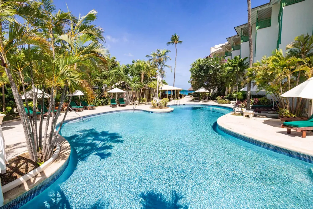Une piscine turquoise bordée de palmiers et de transats sous un ciel bleu.