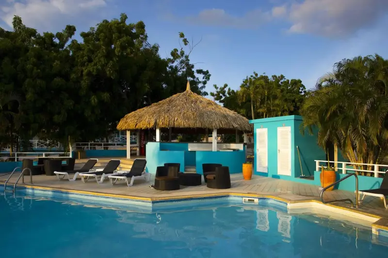 Une piscine turquoise bordée de chaises longues et d'un pavillon de chaume.
