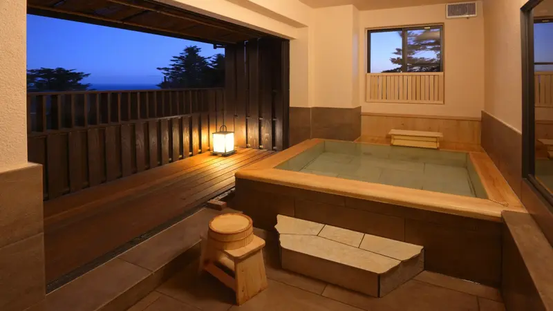 Une baignoire intérieure en bois donne sur une terrasse avec vue sur la mer au crépuscule.