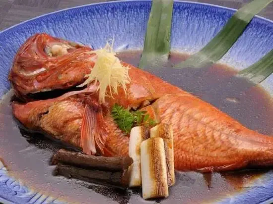 Un poisson entier cuit est présenté dans une sauce sombre, décoré d'herbes et de gingembre râpé.