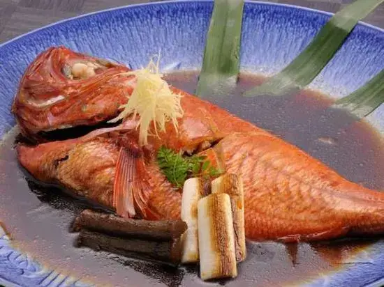 Un poisson entier cuit est présenté dans une sauce sombre, décoré d'herbes et de gingembre râpé.