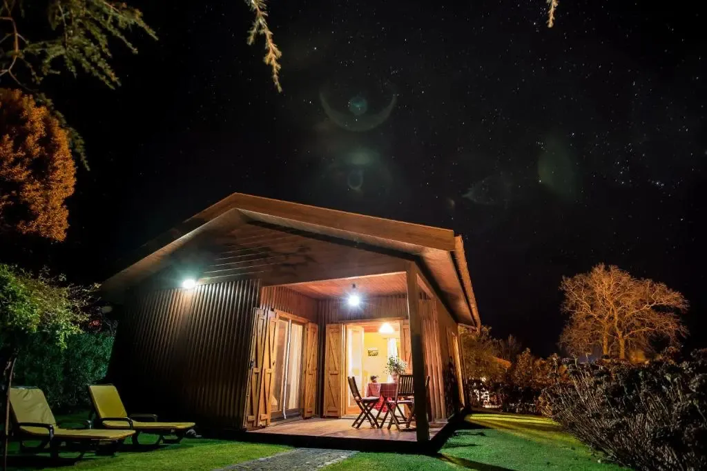 Une petite maison en bois éclairée la nuit avec une terrasse et des chaises longues.