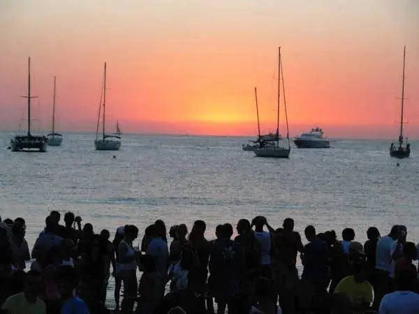 Un groupe de personnes observe un coucher de soleil sur la mer avec des voiliers à l'horizon.