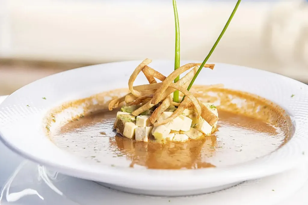 Une soupe crémeuse est servie dans une assiette blanche, garnie de dés de légumes et de fines lamelles croustillantes.
