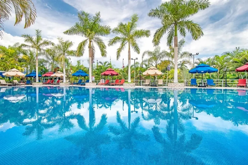 Piscine rectangulaire bordée de transats et de palmiers sous un ciel nuageux.