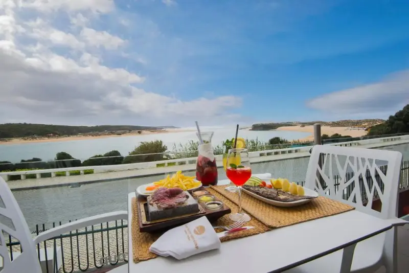 Une table est dressée avec des plats et des boissons sur une terrasse ensoleillée avec vue sur une baie.
