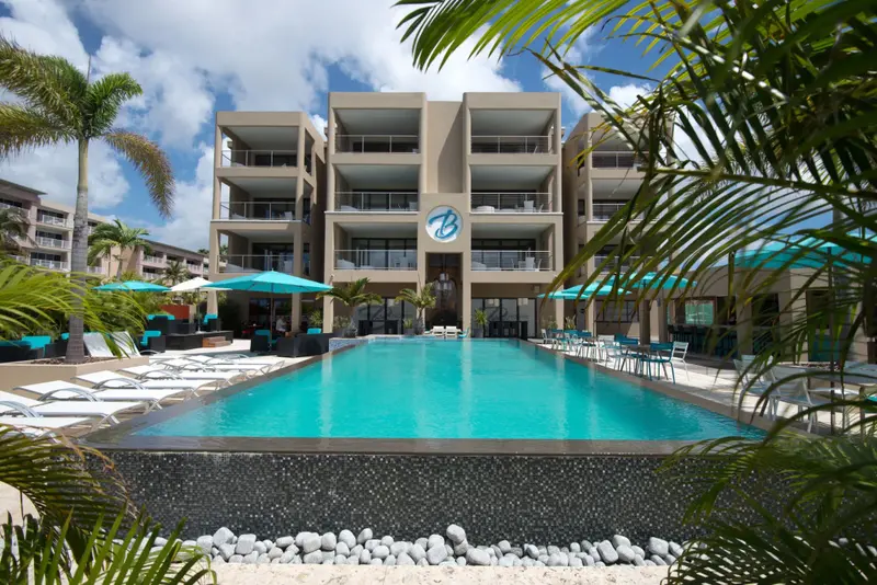 Une piscine turquoise bordée de transats devant un bâtiment beige à balcons.